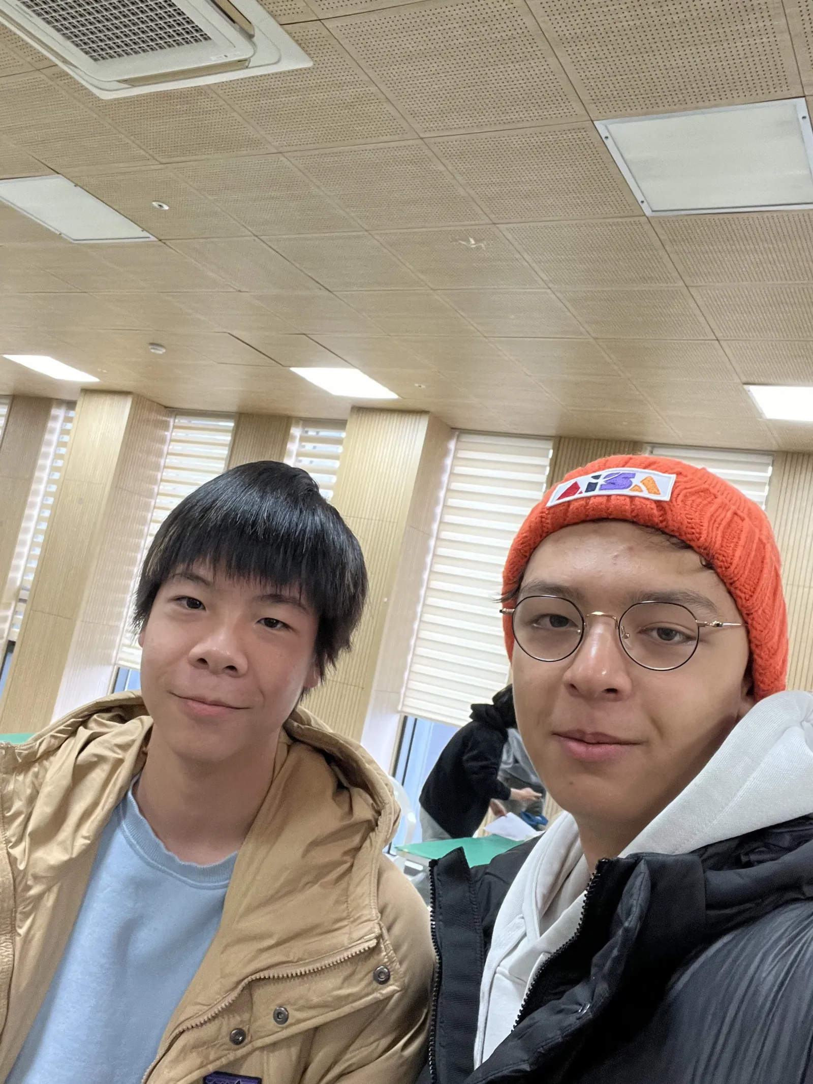 Anders in an orange beanie and glasses taking a selfie with another student in a bright, wood-paneled hall
